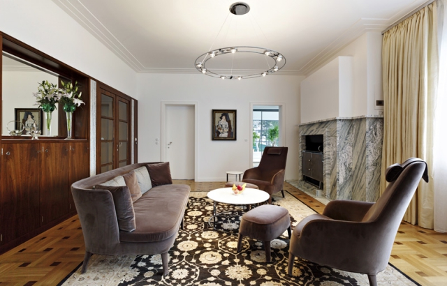 Italská sedací souprava v salonu stylově ladí s historizujícím vybavením interiéru. Mramor cippolino, použitý na krbu, patří k oblíbeným Wiesnerovým materiálům. Charakteristický je i kruhový kovový lustr, vyrobený jako replika.