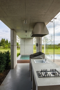 Architekt originálně propojil exteriér domu s interiérem. Betonová stropní deska vybíhající až nad bazén chrání část vodní plochy, ale především venkovní kuchyni, navazující na tu uvnitř domu. Pobyt venku a společné vaření si tak rodina může užívat, jak nejdéle to jde.