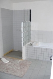Koupelna je obložená bílými a šedými obklady v různých formátech. Luxferová stěna odděluje vanu a sprchový kout