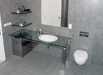 Styl koupelny a její vzdušnost zdůrazňuje skleněný pult se zabudovaným umyvadlem. Jednobarevnou plochu obkladu zdobí pouze vertikálně položené úzké pruhy a listela s drobným dekorem